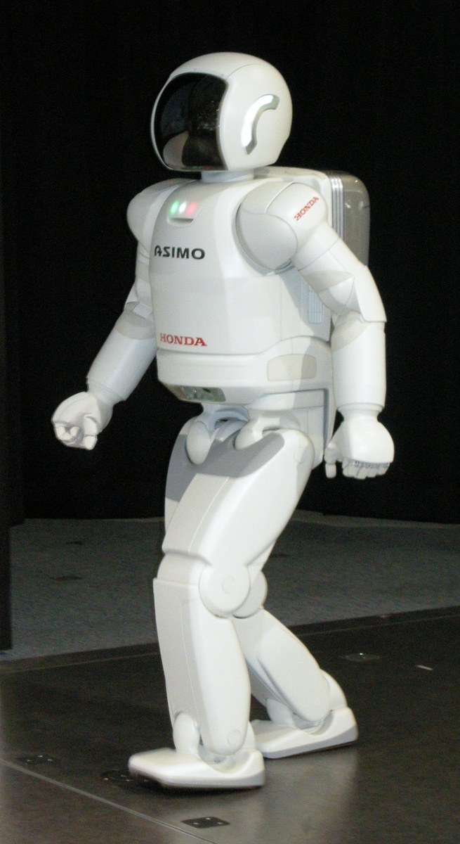 Honda's white ASIMO robot walking across a stage, one foot forward, knees bent, leaning slightly over its front foot.